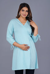 A woman wearing a light blue, long, solid stylish top with 3/4 sleeves, standing against a grey background.